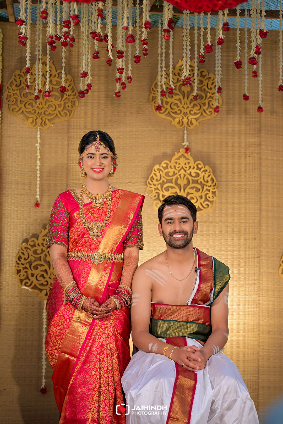 Bride in a pink and gold saree with heavy gold jewelry stands beside a smiling groom in white and gold on a decorated wedding stage.