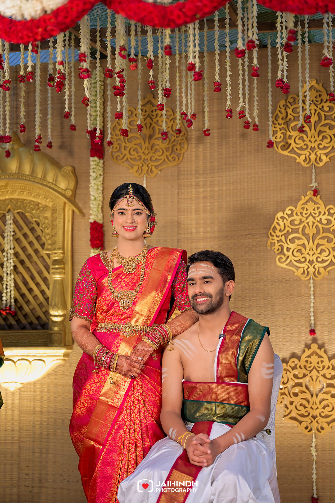 Bride in a pink and gold saree with heavy jewelry stands beside a seated groom in white, on an ornate wedding stage with gold filigree decor and red flowers.