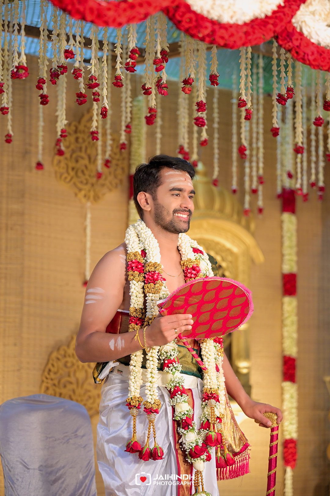 Smiling groom in traditional wedding attire with flower garlands, holding a pink decorative fan under a floral backdrop.