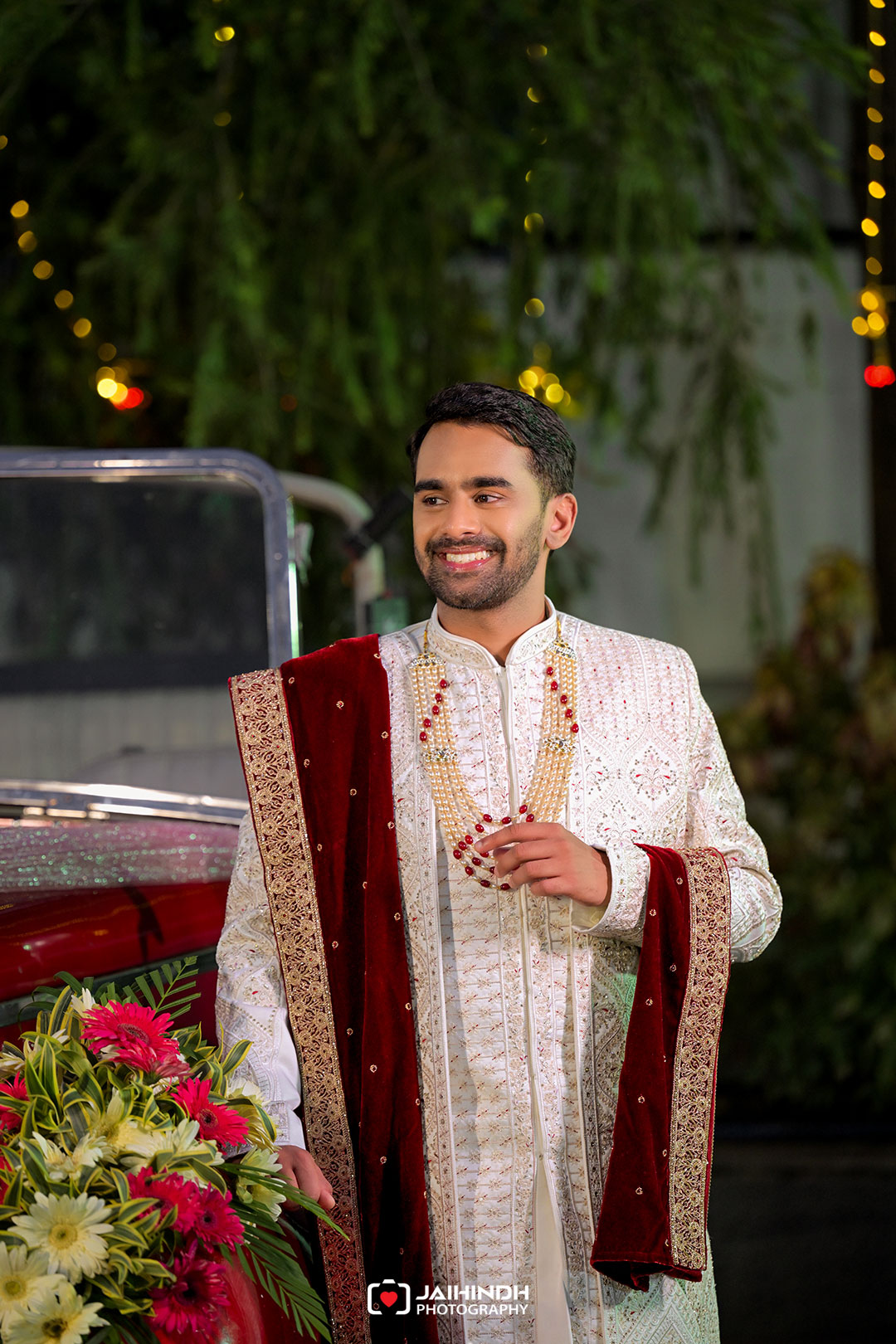 Smiling man in a cream and gold sherwani with a maroon velvet stole, posing beside a vibrant flower arrangement at a night wedding backdrop.