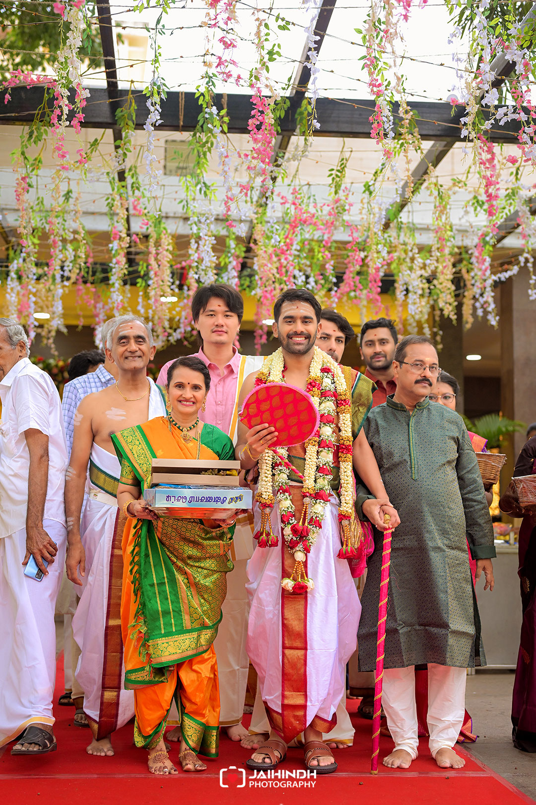 Groom in white and red ceremonial attire with flower garlands poses with family and friends under a canopy of hanging pink and white blossoms.