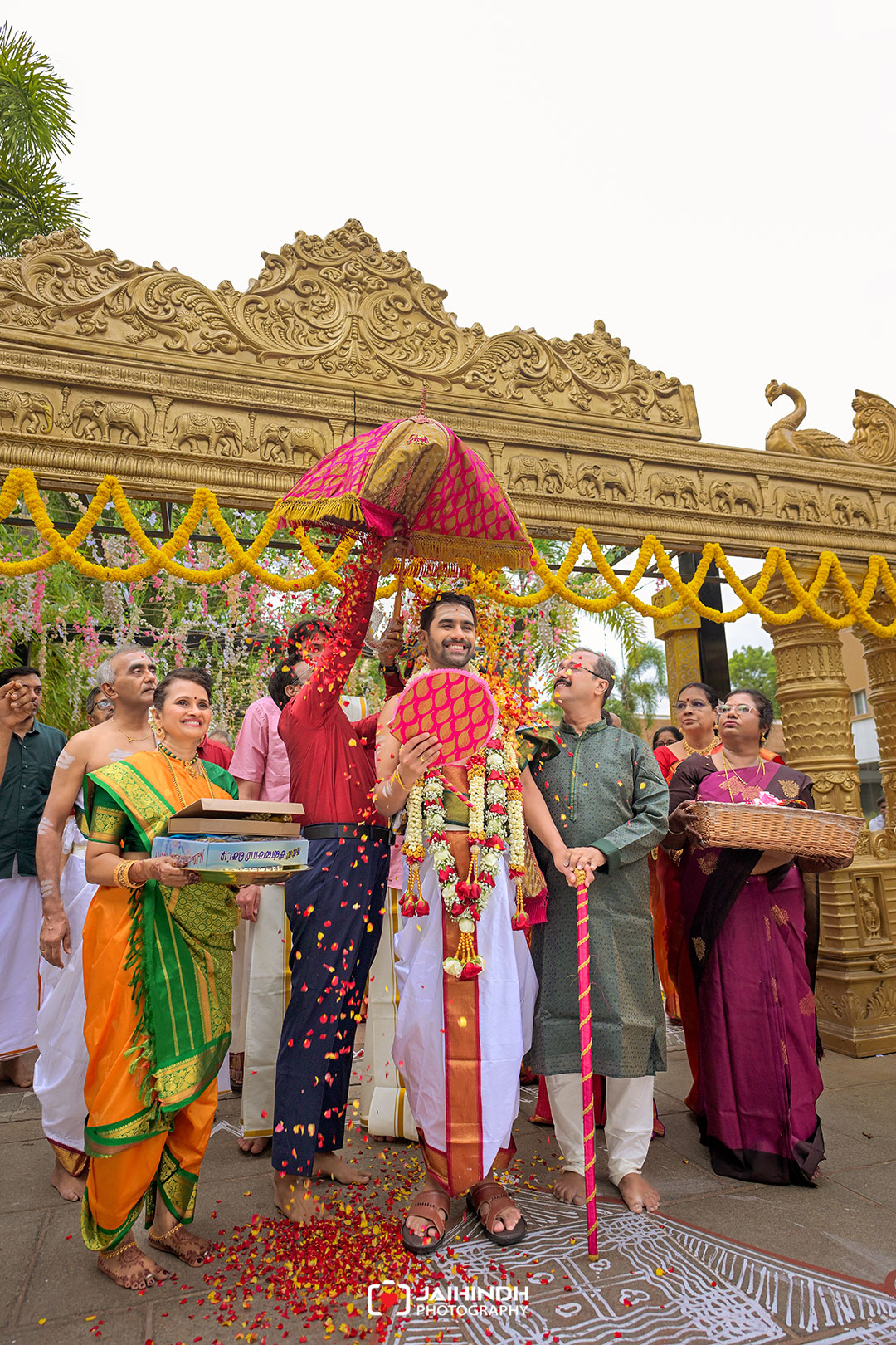 Groom under a pink ceremonial umbrella surrounded by family during a colorful Indian wedding in front of a carved gold temple arch, petals falling around them.