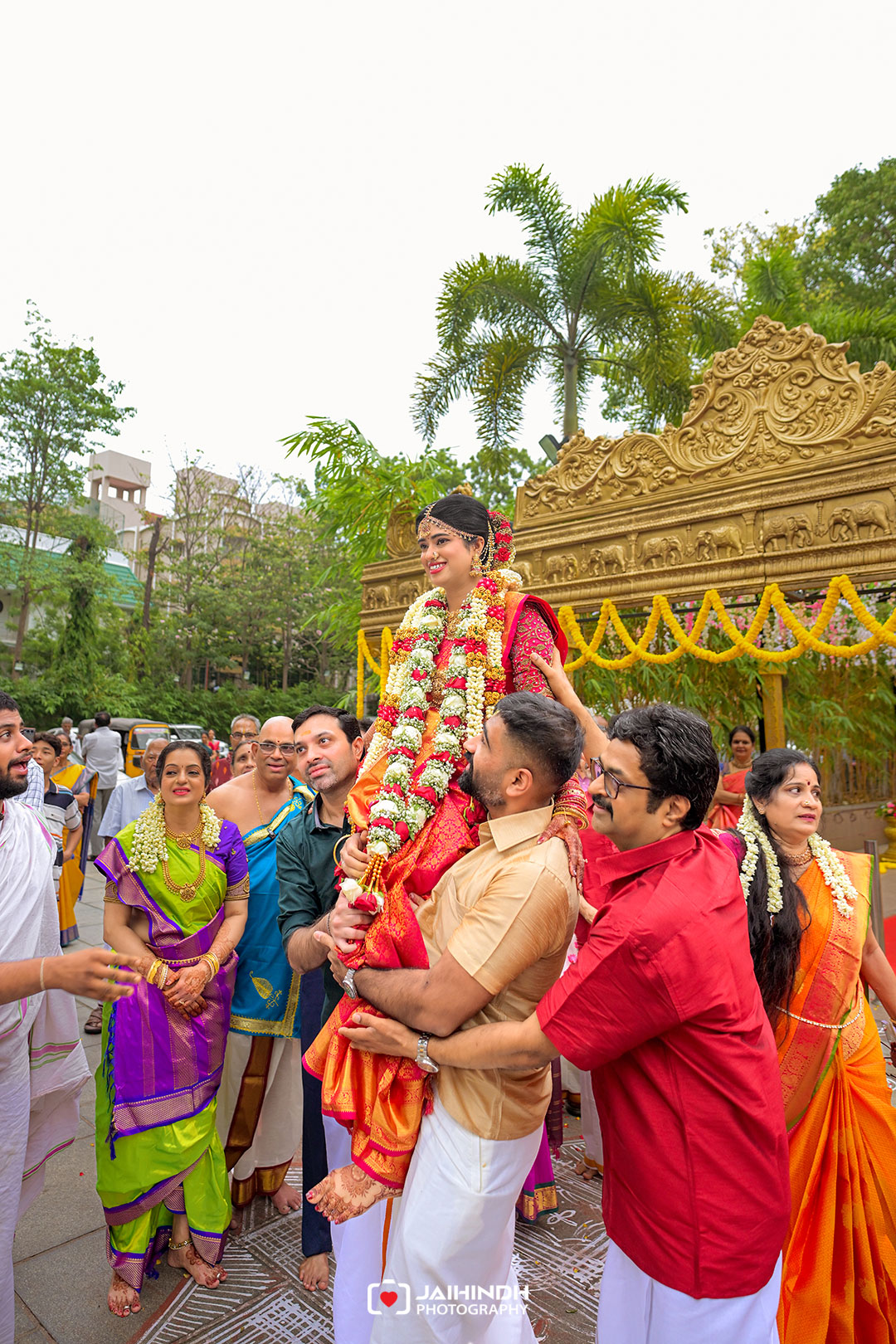 Bride in a red lehenga lifted on shoulders during a Hindu wedding, surrounded by family in bright sarees and a carved gold backdrop.