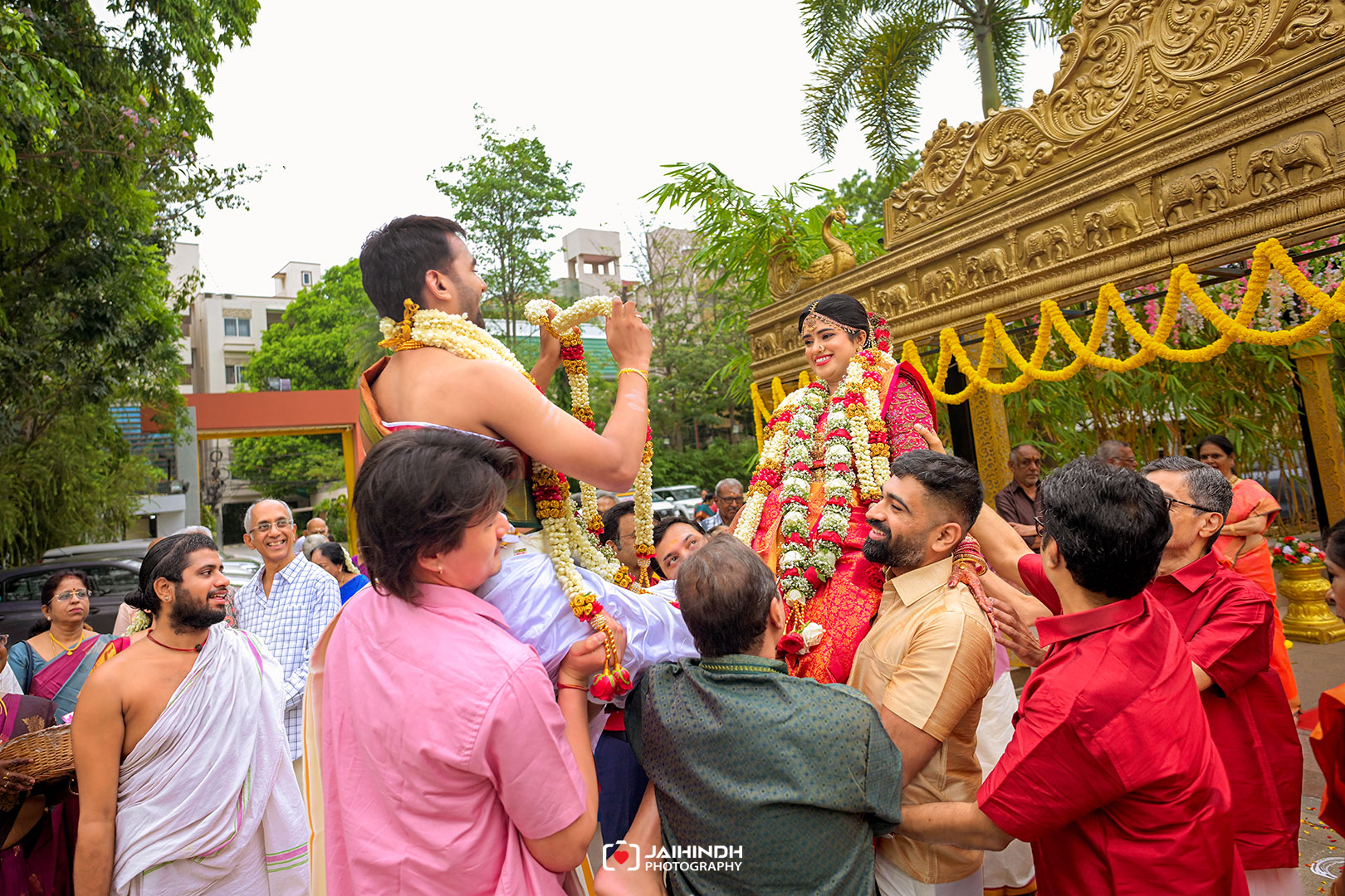 Bride in a red lehenga with flower garlands being carried by friends at a Hindu wedding beside an ornate gold decorative backdrop.