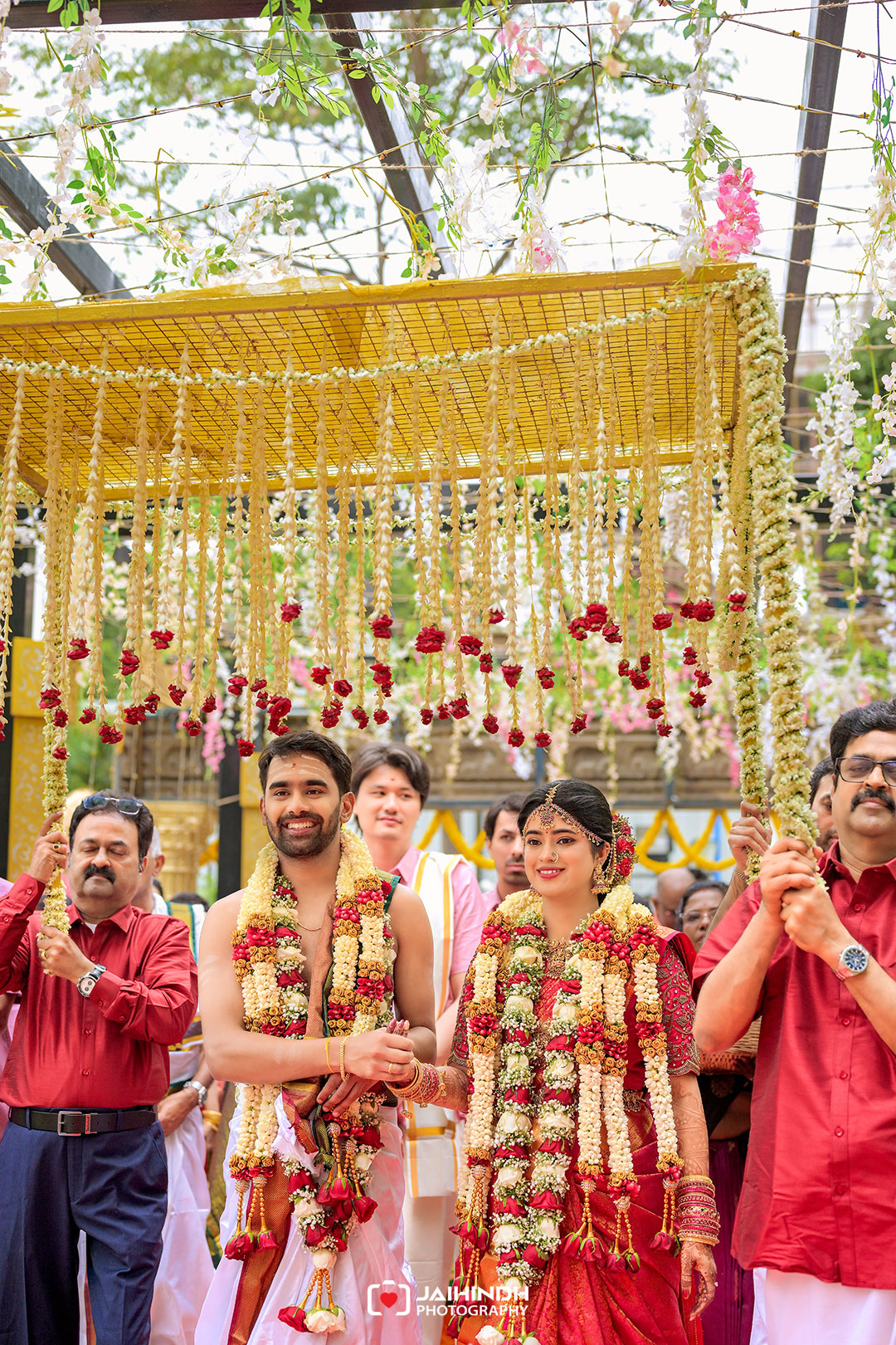 Bride and groom in traditional attire with flower garlands, walking under a canopy of hanging floral strings at a wedding.