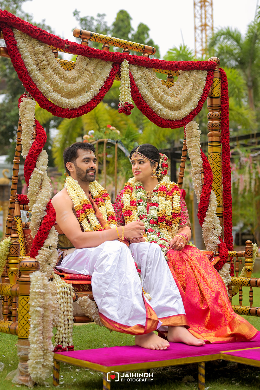 Bride and groom in traditional Indian attire sit on a flower-decorated swing, wearing garlands outdoors at their wedding.