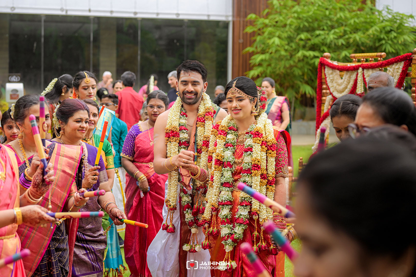 Bride and groom wearing flower garlands walk through a festive crowd of relatives in colorful sarees at an outdoor Indian wedding procession.