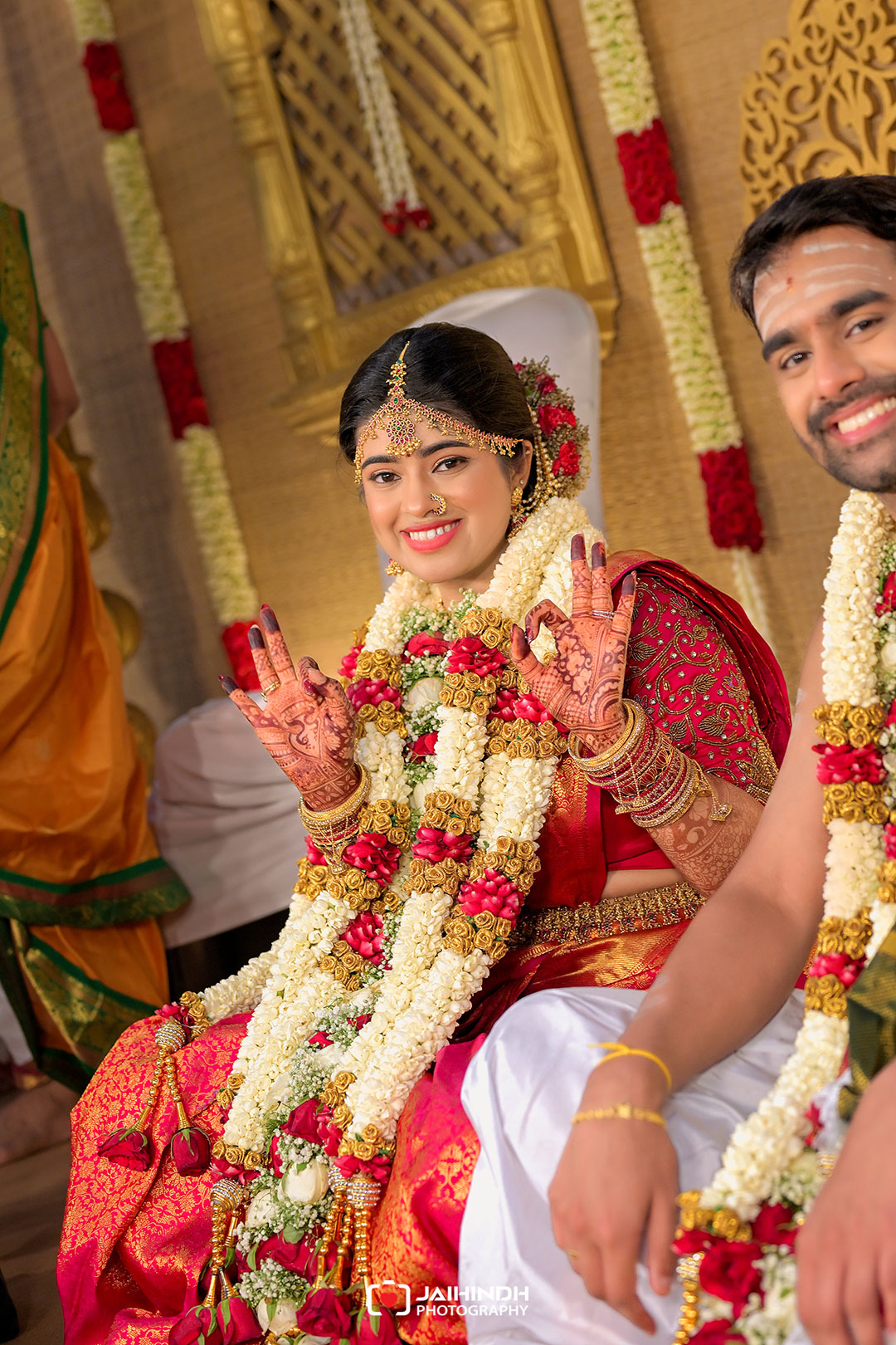 Bride in red bridal saree with elaborate gold jewelry and white flower garlands showing her hands in a blessing/hello gesture at a wedding ceremony.