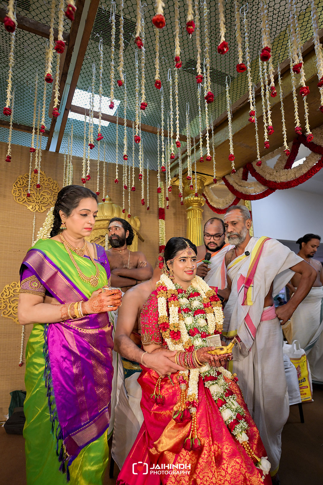 Bride in a red and gold lehenga with flower garlands, surrounded by family during a traditional Indian wedding ceremony under hanging floral decorations.