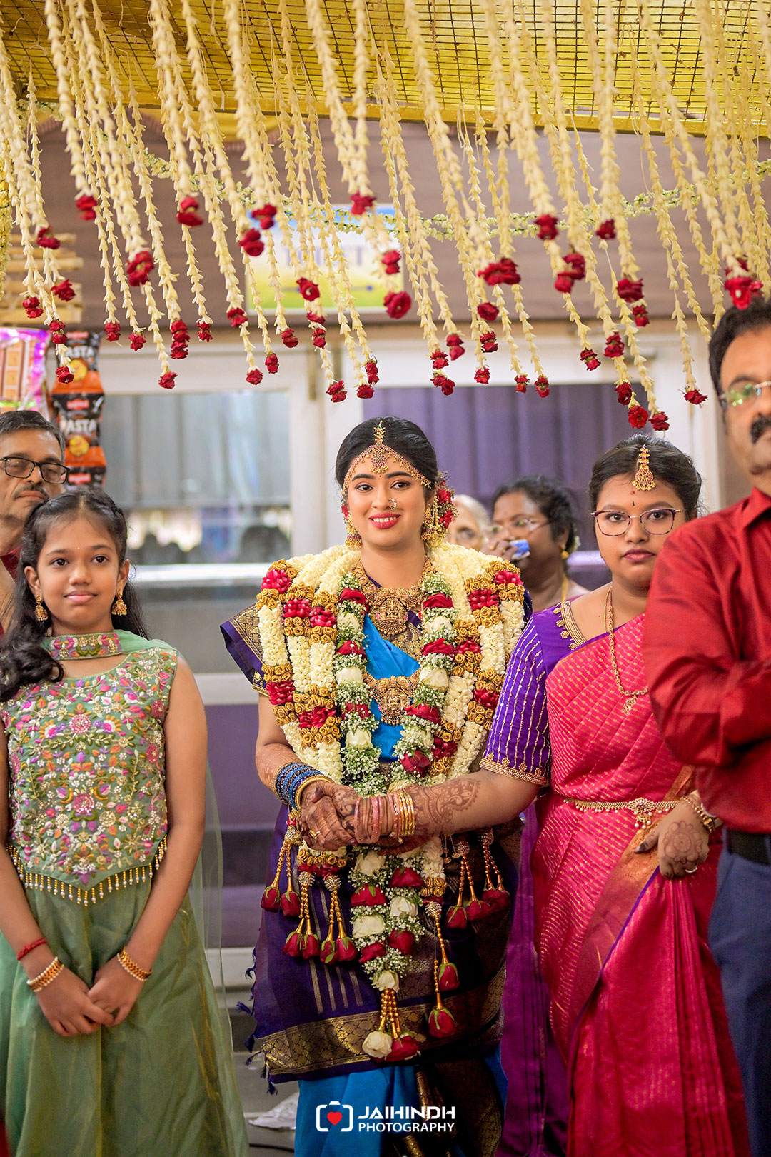 Bride in a blue and purple sari wearing elaborate flower garlands and traditional jewelry, standing with family under hanging floral decorations at a festive wedding scene.