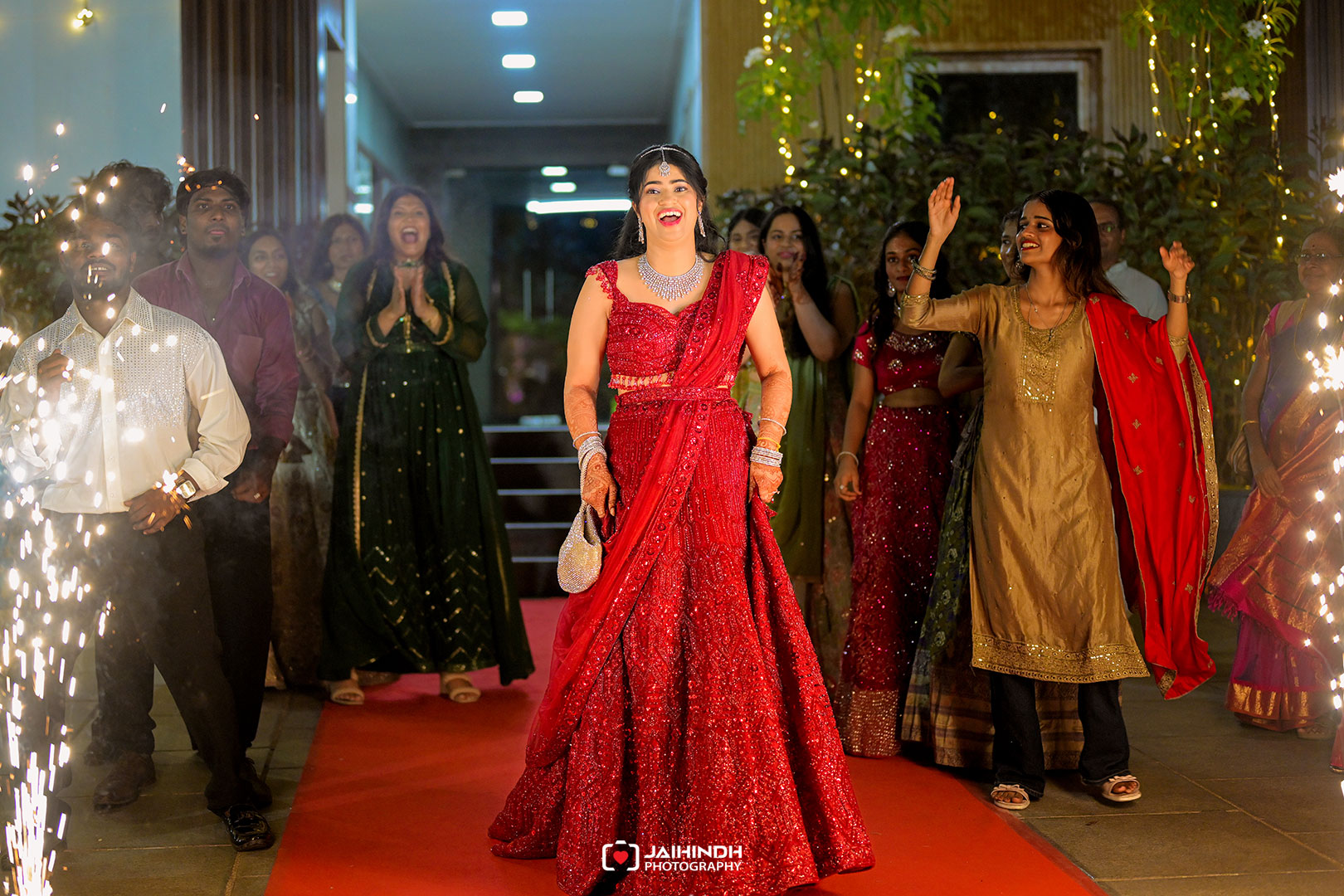 Woman in a red embellished lehenga smiles on a red carpet as friends cheer and sparkler lights glow nearby.