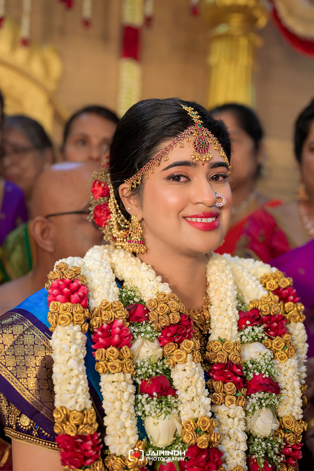 Bride in blue and gold saree with elaborate gold jewelry and white flower garlands, smiling at her wedding.