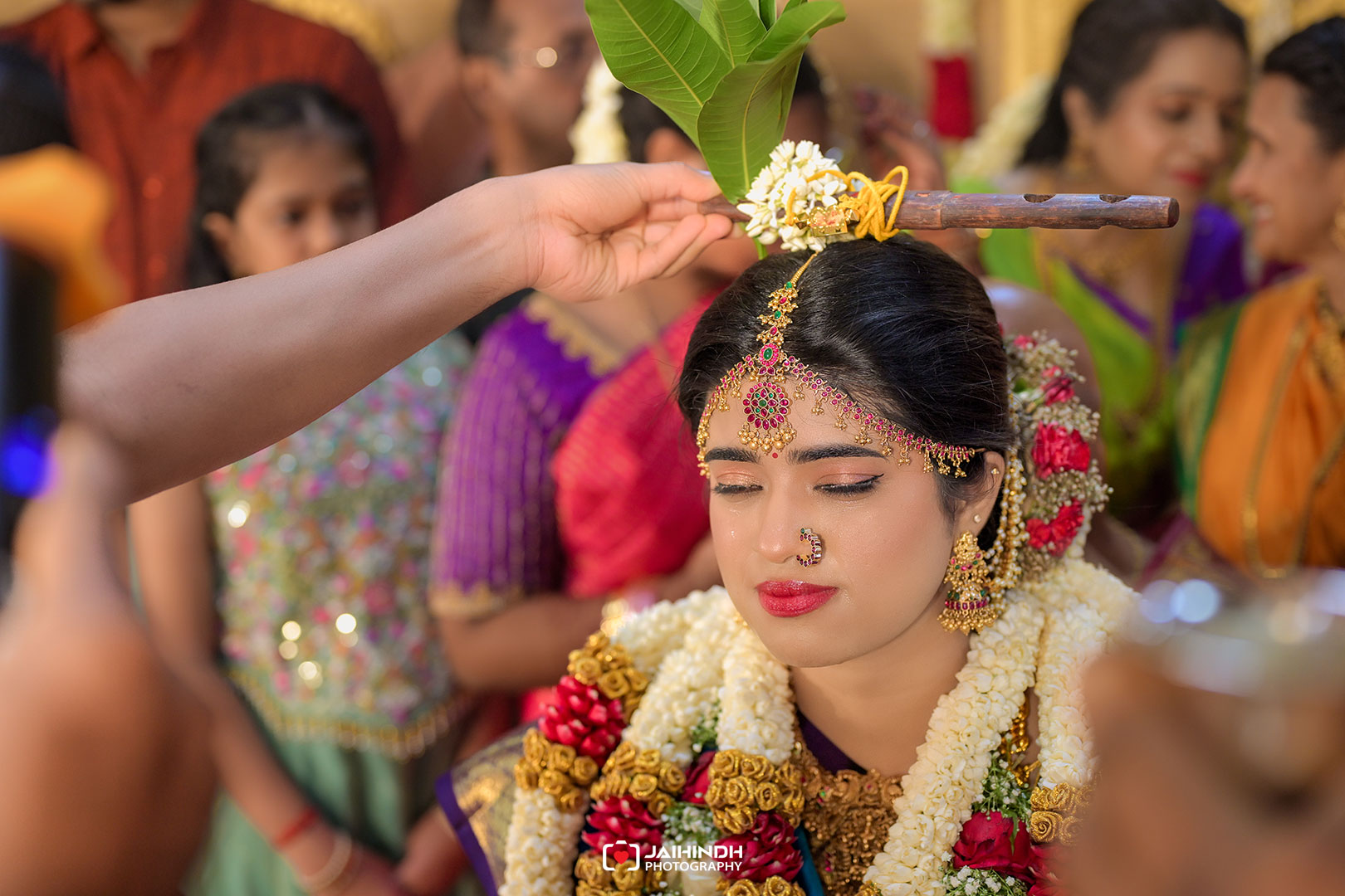 Bride in ornate gold jewelry and garlands, eyes closed as a ceremonial flower-adorned staff is placed on her head by a helper, surrounded by family in colorful attire.