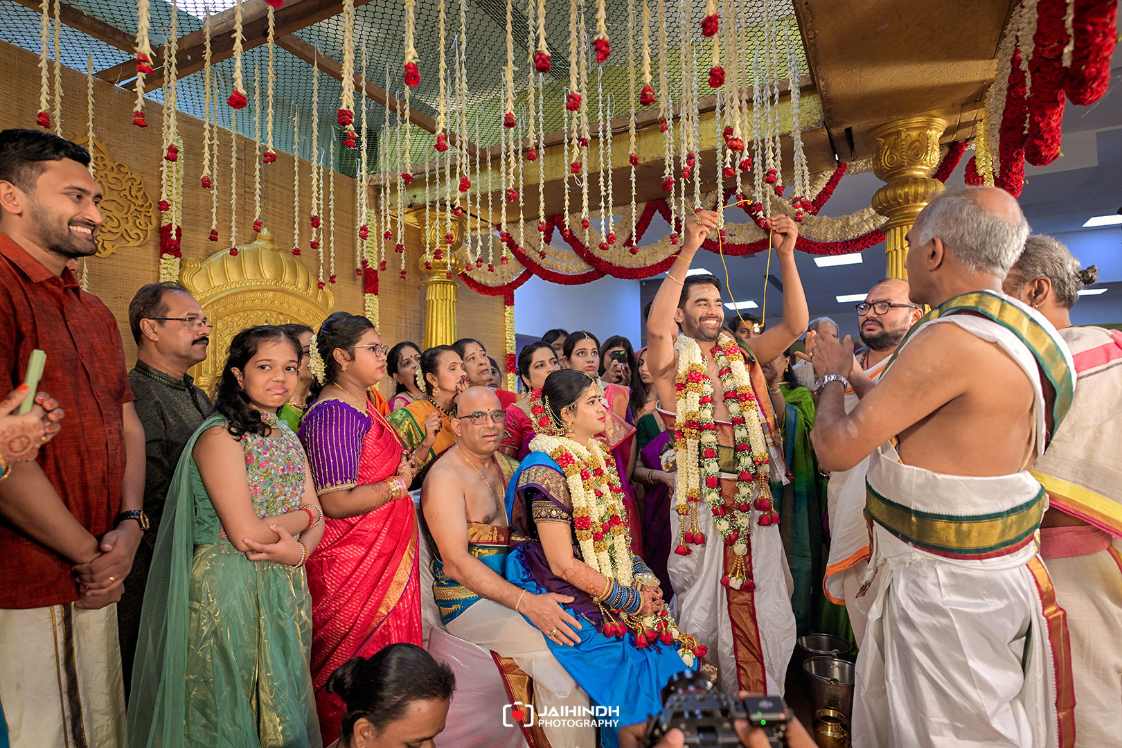 Bride and groom seated center, wearing flower garlands, surrounded by family in colorful sarees and dhotis at a Hindu wedding under hanging floral decorations.