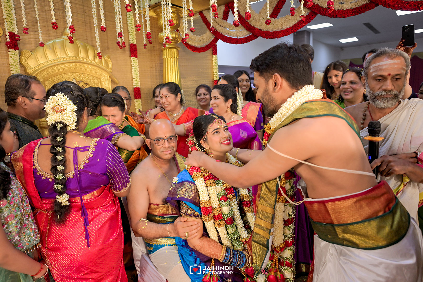 Bride in a blue sari sits smiling as her groom places a garland during a traditional Indian wedding surrounded by family in colorful sarees and decorations.