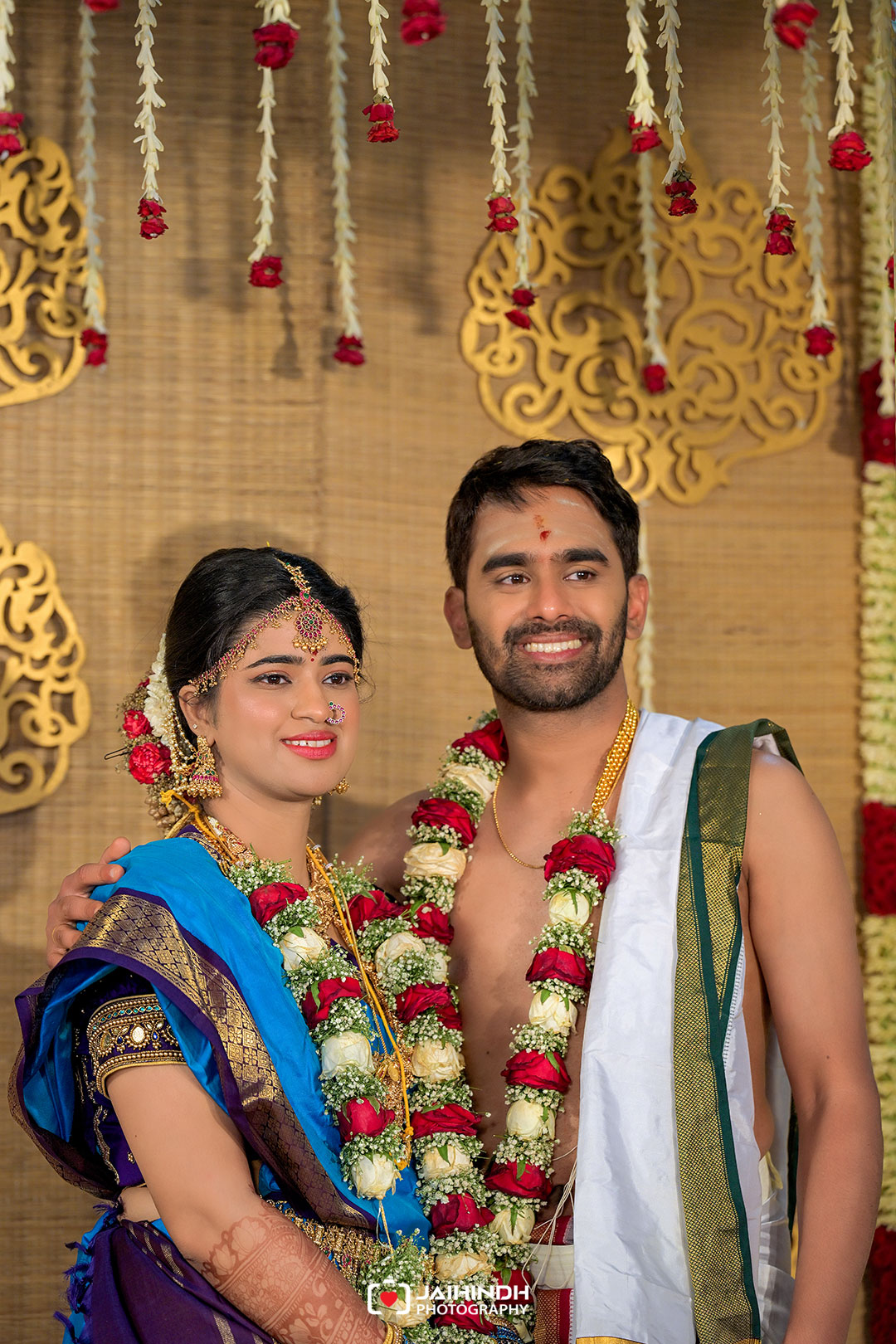 Bride and groom pose together at their wedding, both smiling, wearing garlands and traditional Indian attire with a decorative backdrop behind them.