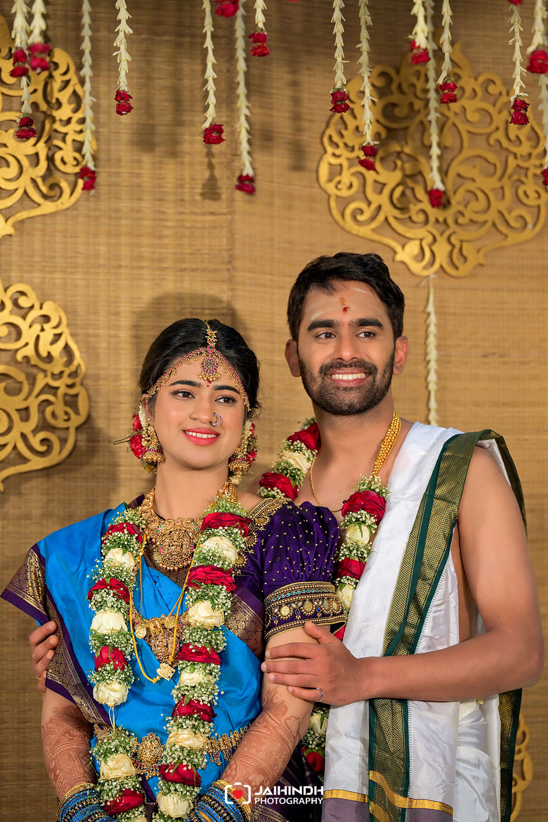 Bride and groom in traditional attire smiling, wearing flower garlands, posing together at a festive ceremony backdrop