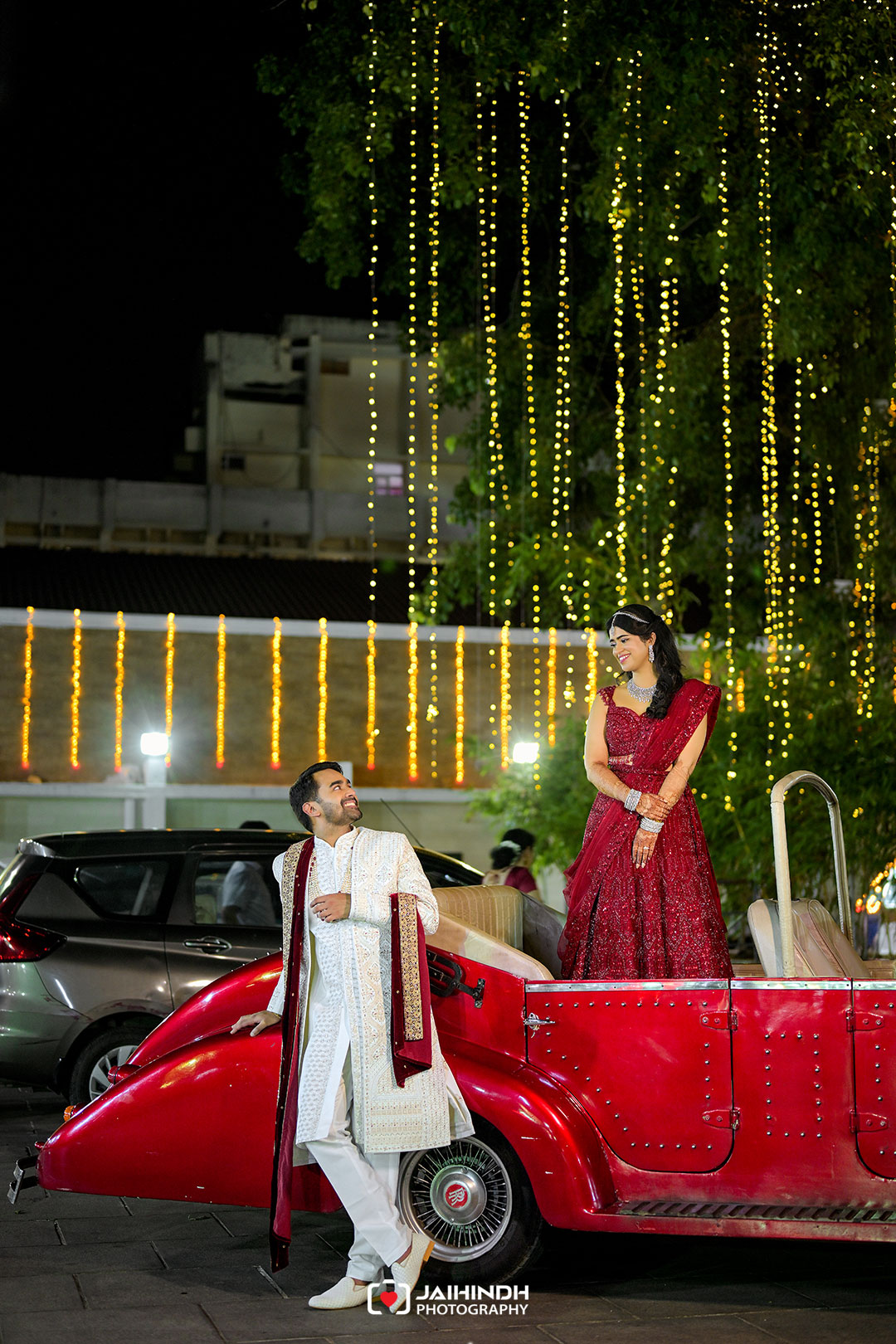 Bride in a red lehenga stands in a vintage red car while groom in cream sherwani smiles at her, backdrop of hanging fairy lights at night.