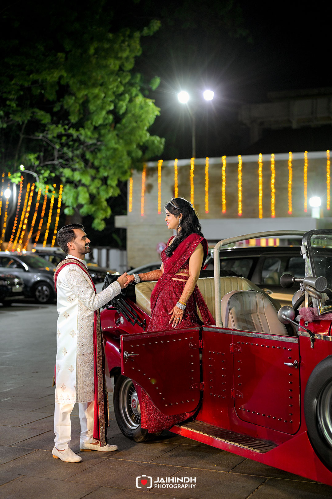 A couple in traditional Indian wedding attire shake hands beside a red vintage car at night, string lights in the background.