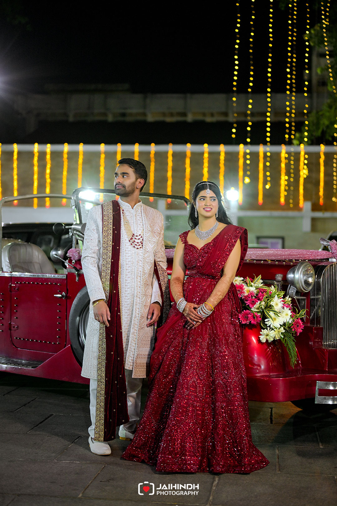Couple in traditional wedding attire posing beside a vintage red car at night with string lights in the background