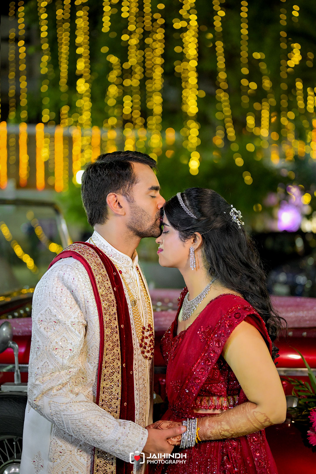Bride and groom in ornate Indian wedding attire share a kiss at night, golden fairy lights creating a bokeh backdrop.