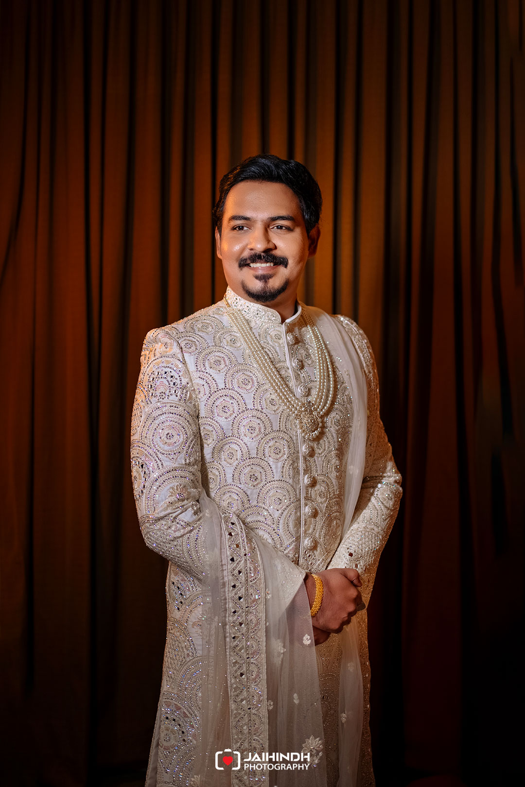 Portrait of a man in a white, heavily embroidered sherwani with pearl necklaces, standing with hands clasped against a warm brown curtain backdrop.