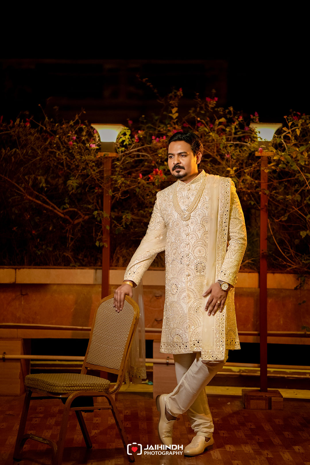 Man in an ivory, heavily embroidered sherwani standing beside a chair on a night-lit terrace garden.