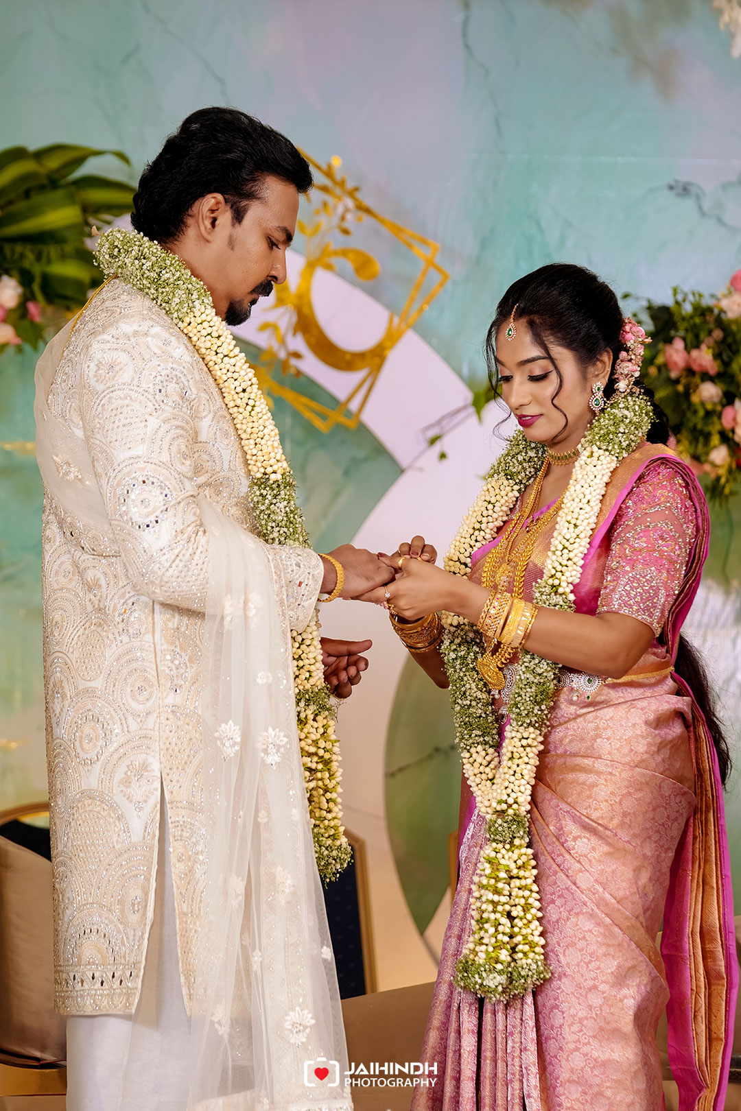Bride and groom exchange rings during an indian wedding, wearing jasmine garlands and ornate traditional attire on a colorful backdrop.