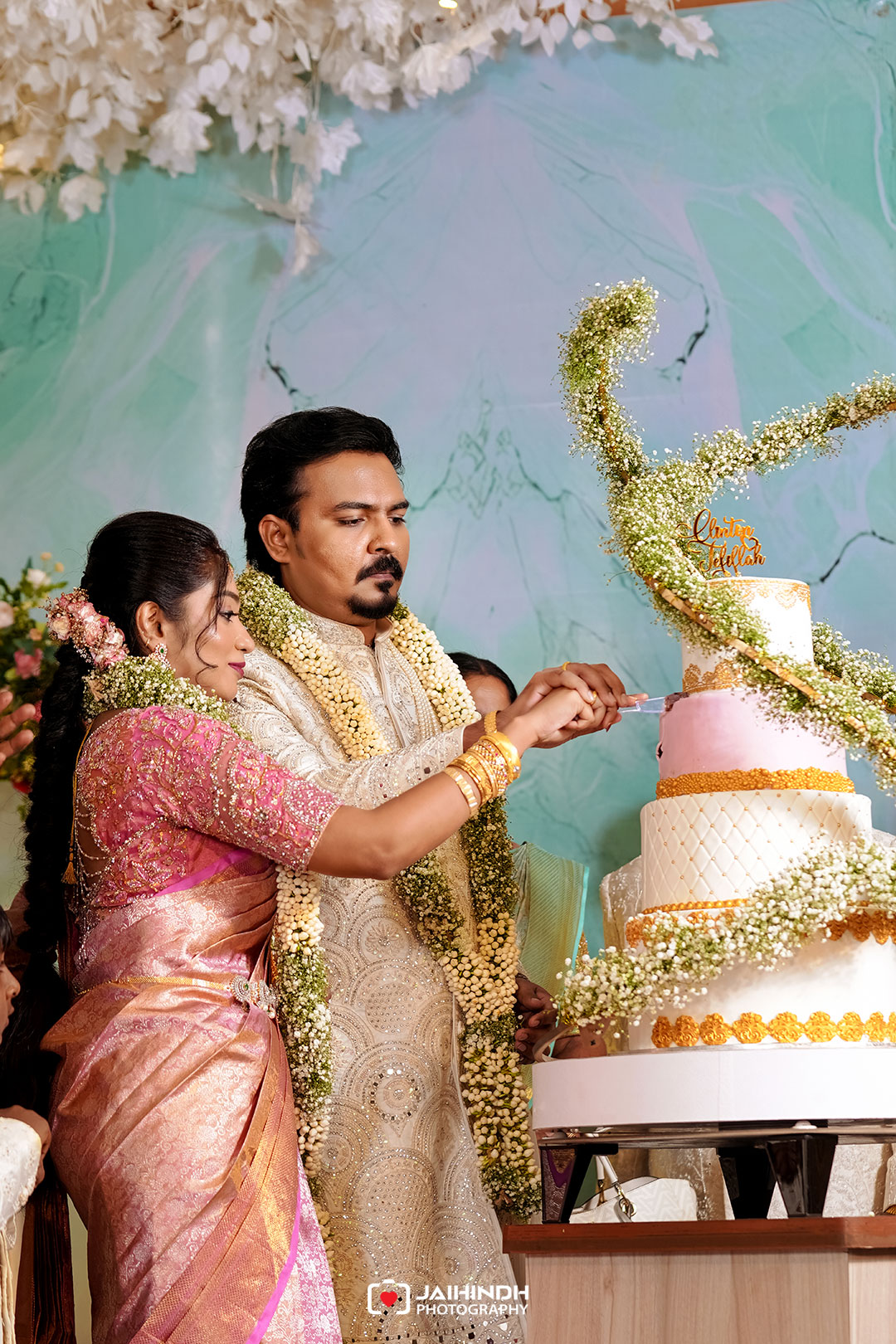 Bride and groom in ornate ethnic outfits cutting a multi-tier wedding cake together, wearing flower garlands.