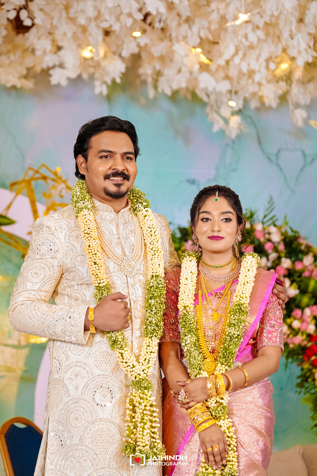 Indian bride and groom stand together in wedding attire, wearing flower garlands and ornate jewelry, posing against a floral backdrop.