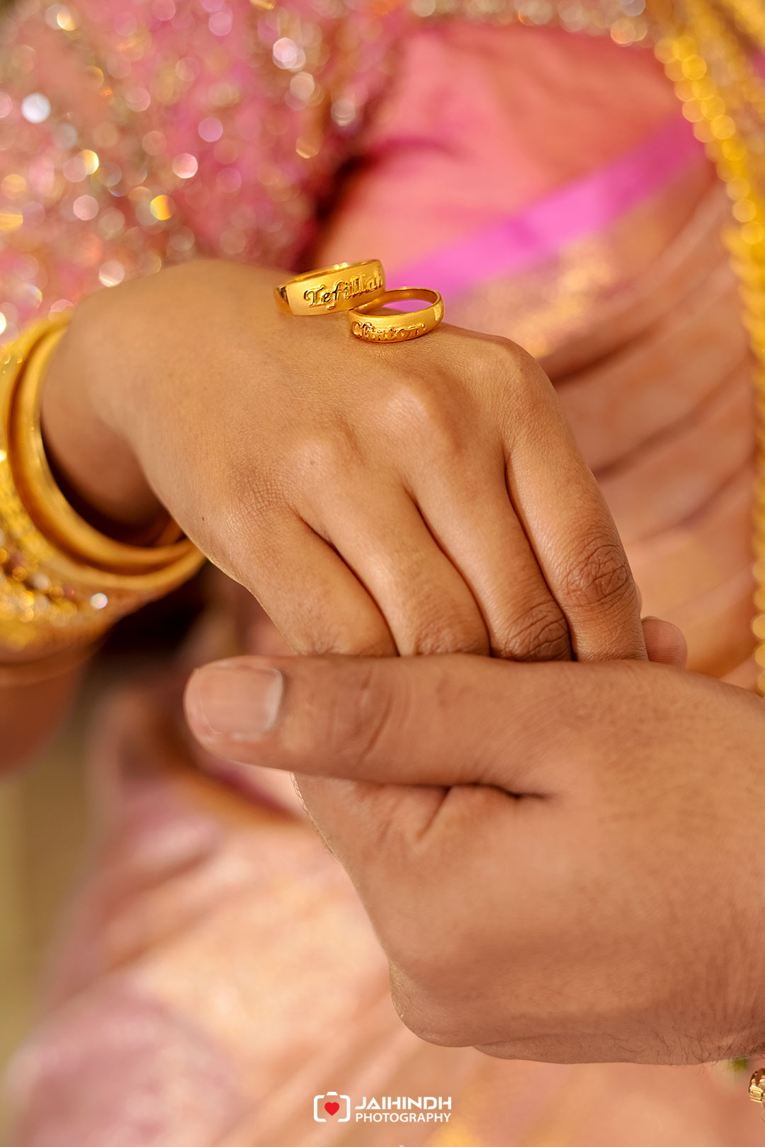 Close-up of a hand wearing gold rings, one engraved, with pink glittery traditional attire in the background.