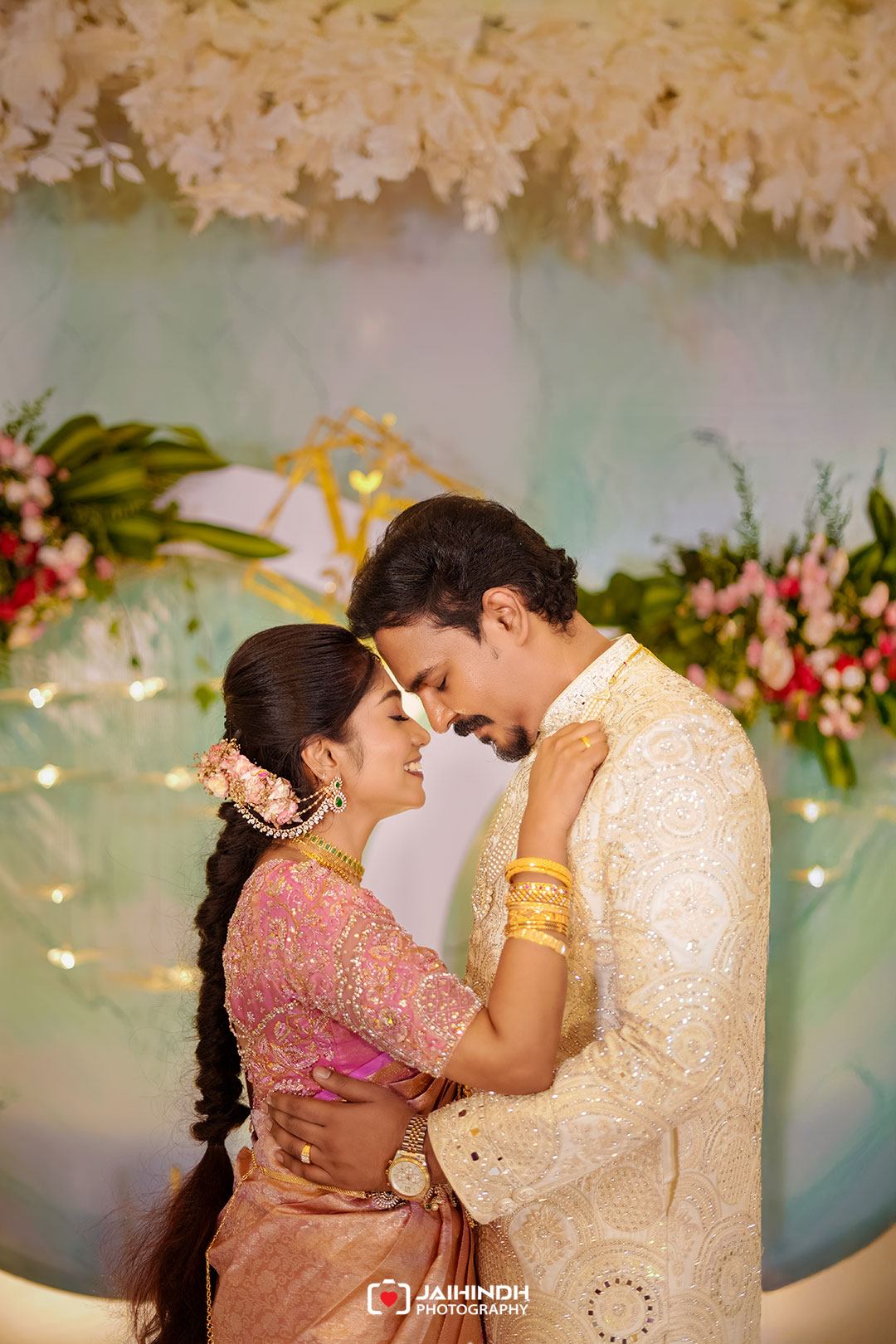 Couple in ornate traditional indian attire embrace, foreheads touching and smiling against a floral backdrop.