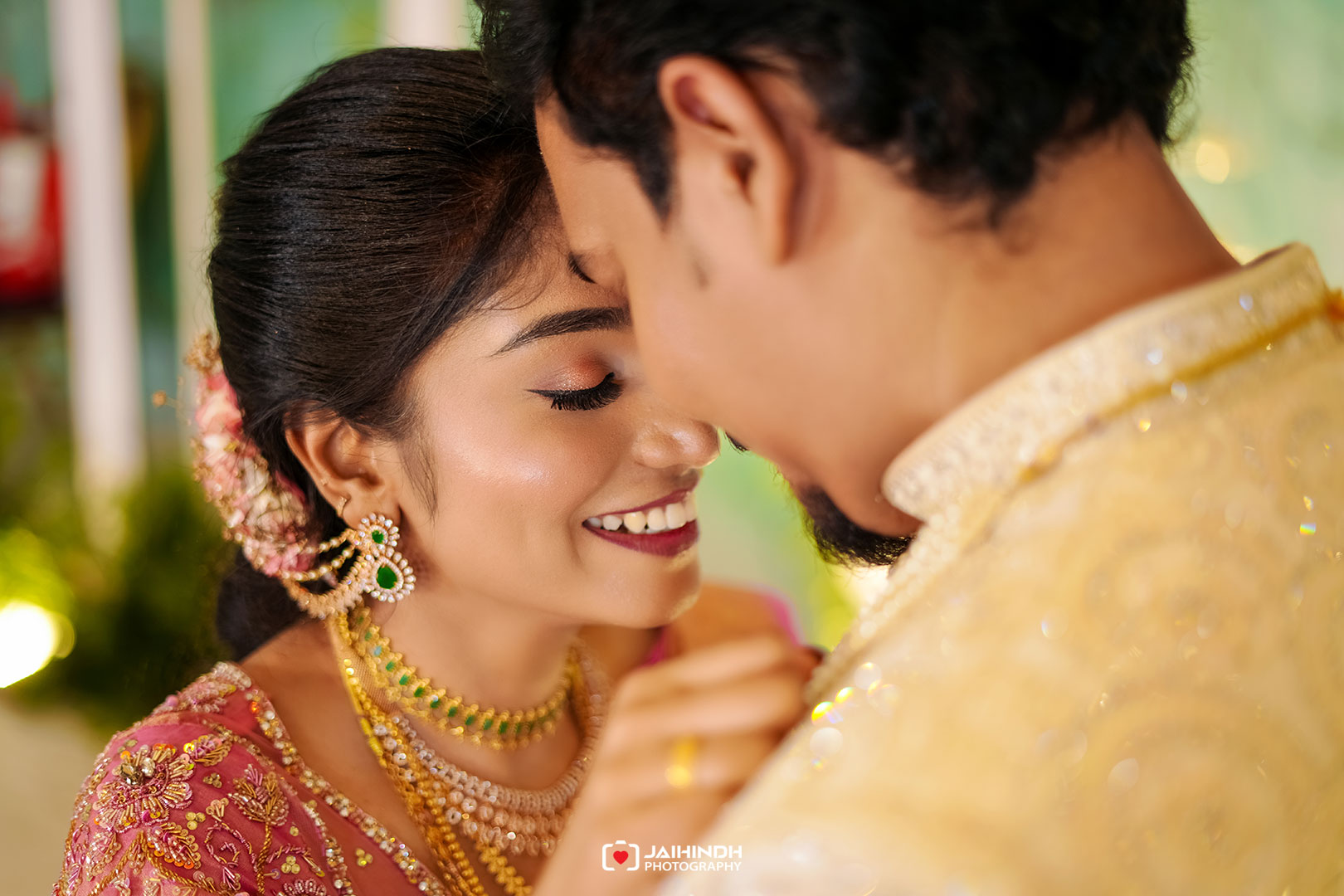 Close-up of a smiling couple sharing a tender moment, bride in pink traditional attire and jewelry, forehead-to-forehead kiss implied by proximity.