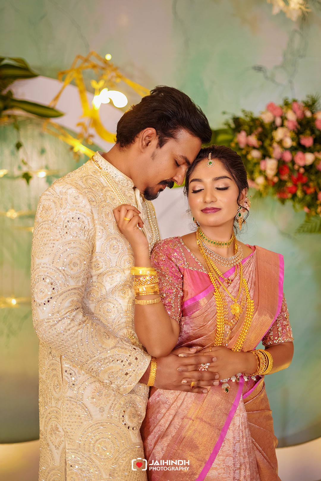 Couple in ornate wedding attire embrace, heads bowed together, amid a floral backdrop and warm lighting.