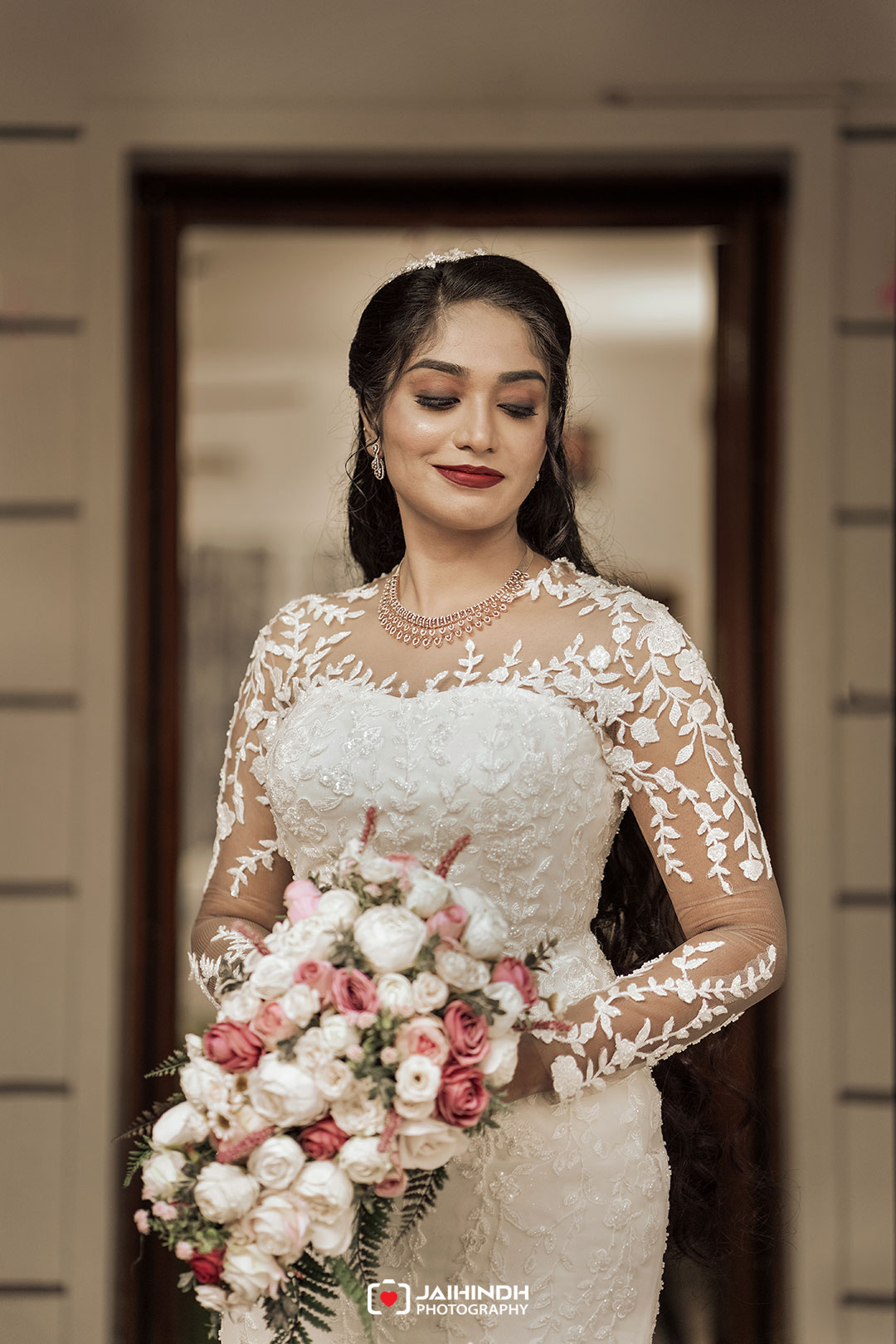 Bride in a lace wedding gown holding a pastel bouquet, standing in a doorway with her eyes closed and a soft smile.