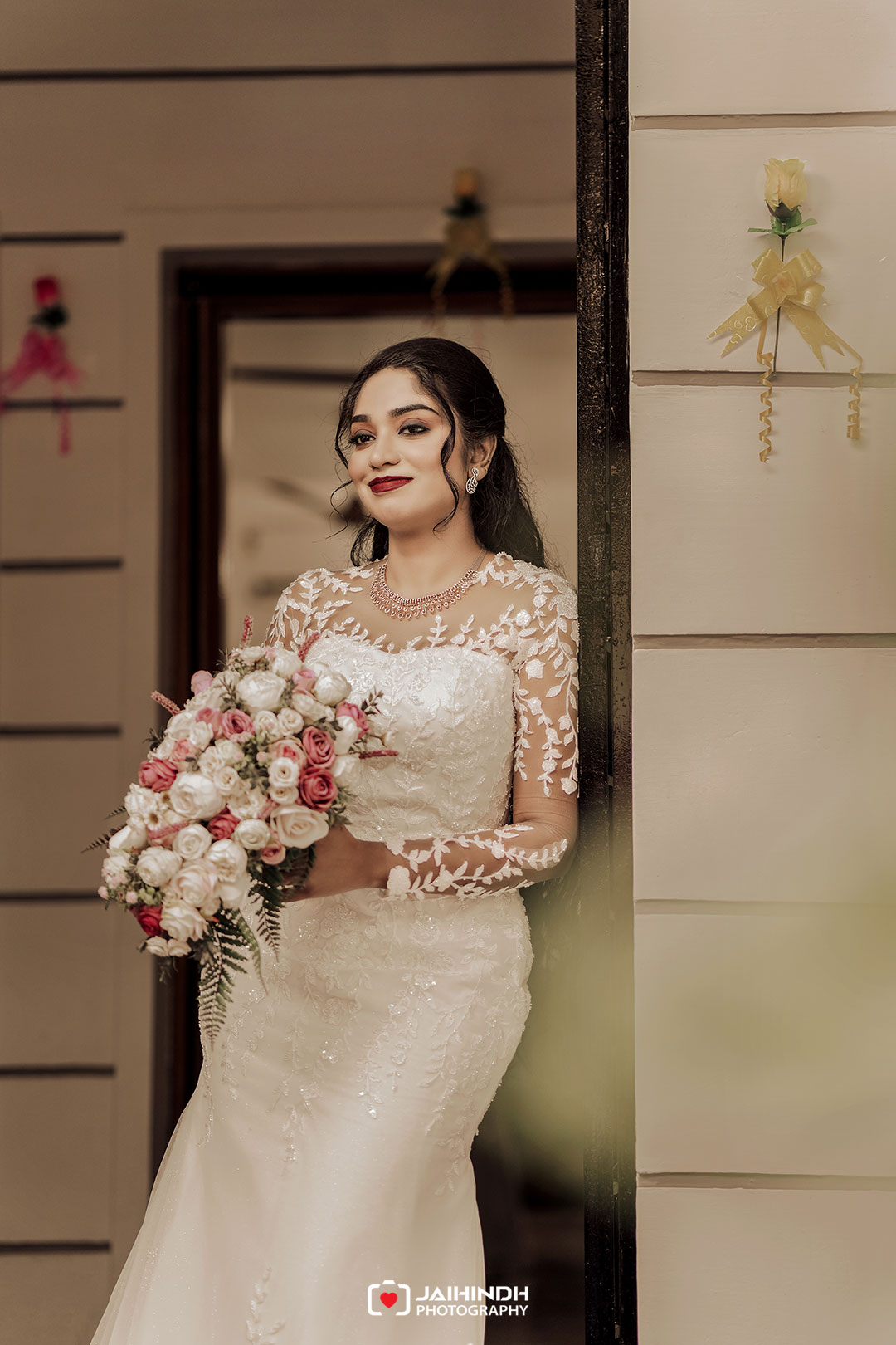 Bride in a lacy, long-sleeve wedding gown holding a pink and white bouquet, standing in a doorway with a soft smile.