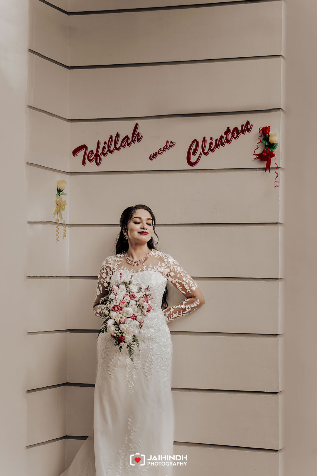 Bride in a white lace wedding dress holding a bouquet of pink and white flowers, standing beside a wall with wedding vows sign.