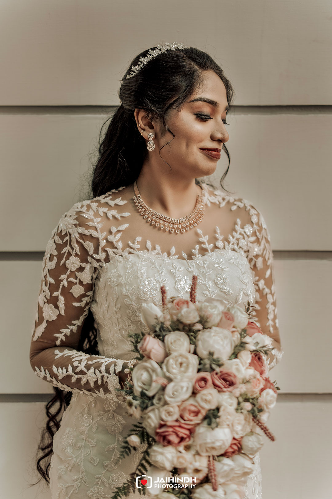 Bride in a lace wedding gown holding a pastel rose bouquet, wearing a pearl necklace and tiara, smiling in profile.