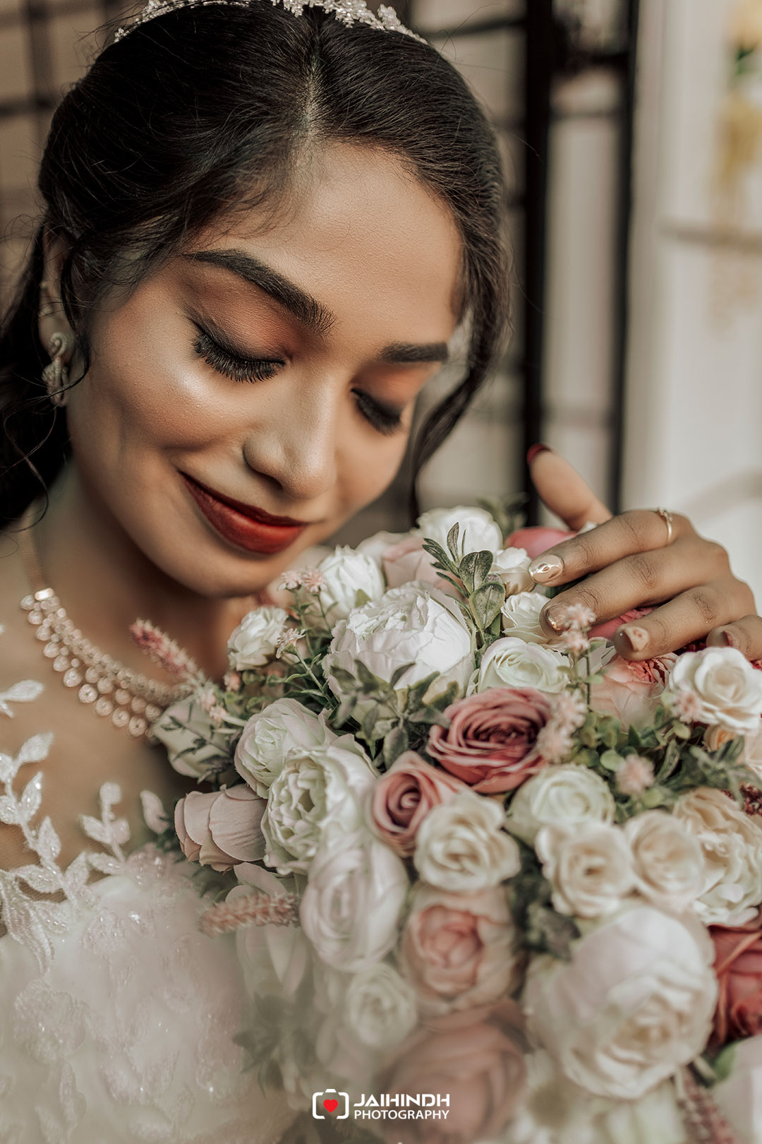 Close-up of a bride with a white lace dress and pearl necklace, smiling with eyes closed while holding a bouquet of white and blush roses.