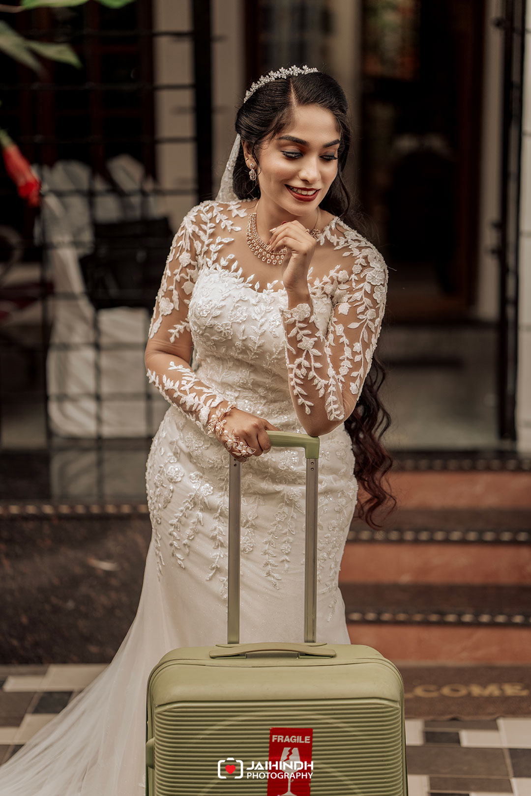 Bride in a lace wedding gown with a tiara and veil, smiling as she holds a green wheeled suitcase labeled fragile outside a building.