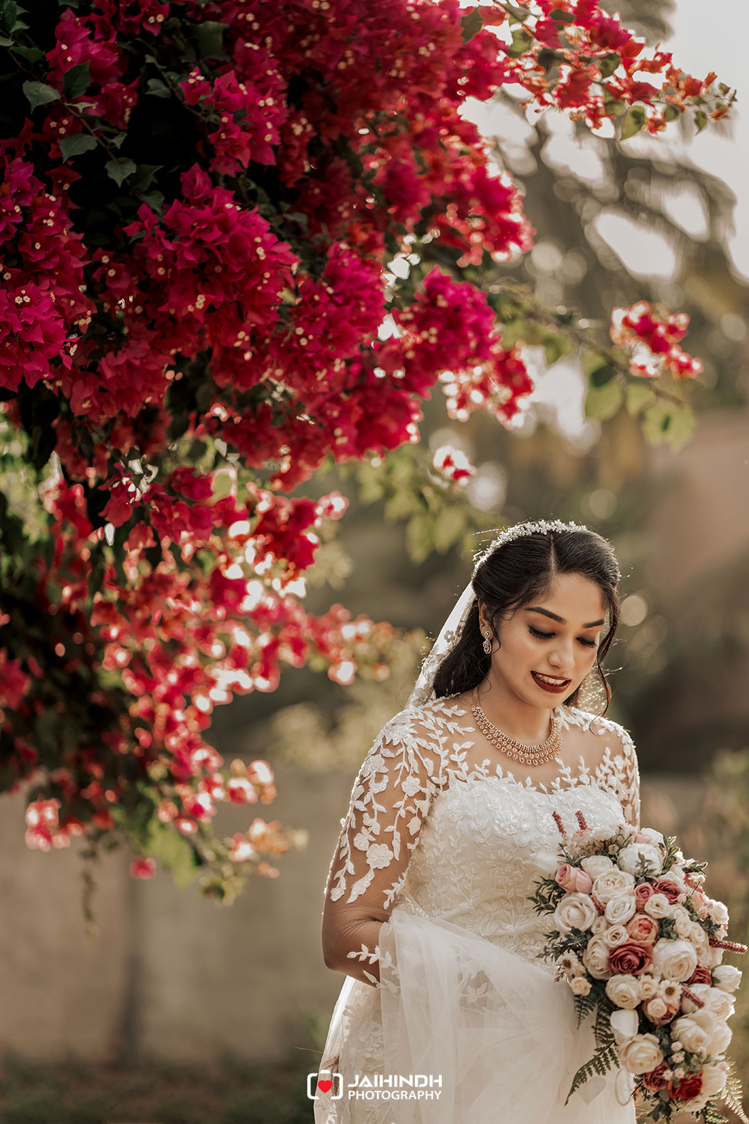 Bride in a lace wedding gown and veil holds a bouquet of pale roses beneath a cascade of magenta bougainvillea flowers outdoors.