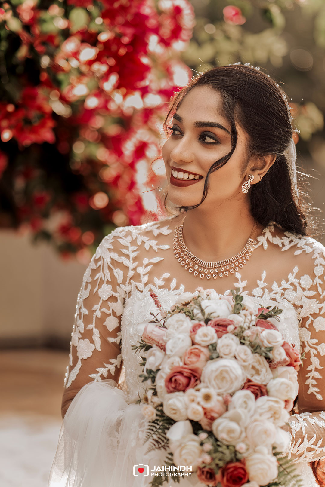 Bride in a lace wedding dress holding a bouquet of blush and ivory roses, smiling with dark lipstick, jewelry, and a floral background behind her.
