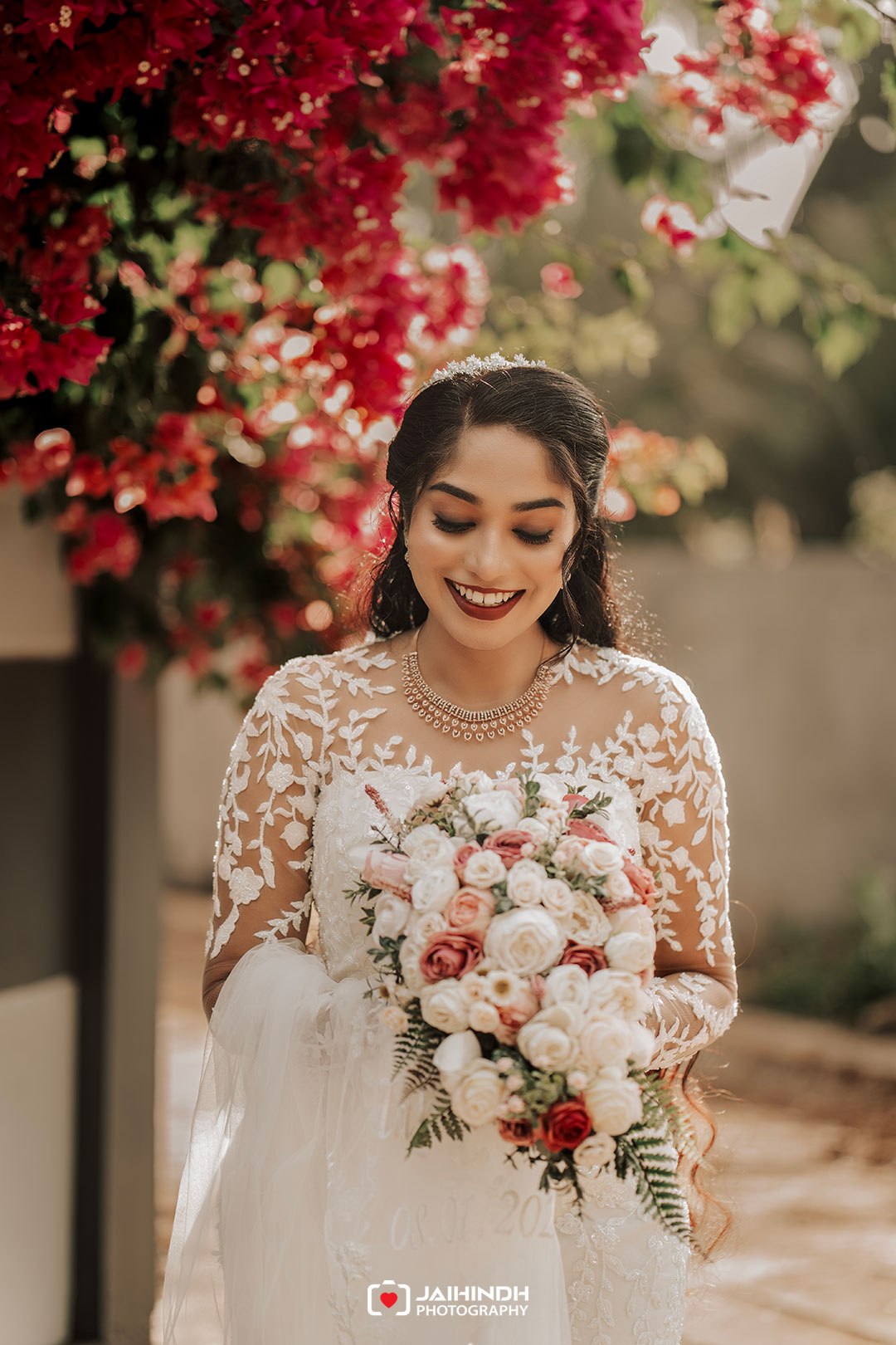 Bride in a lace wedding dress holding a pink-and-white bouquet, smiling as red flowers frame her.
