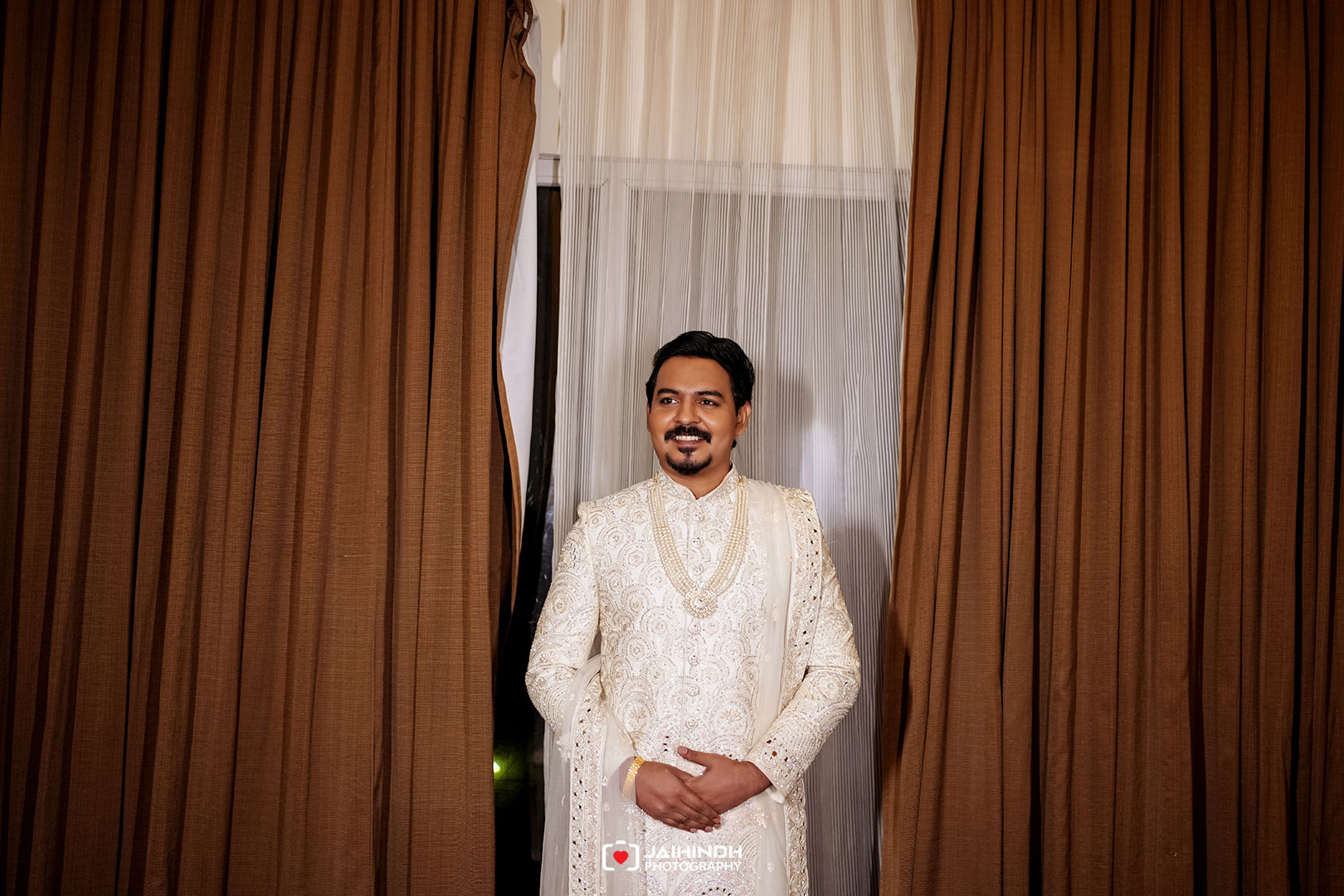 Man in an ornate white sherwani stands between brown curtains, smiling with hands clasped in front.