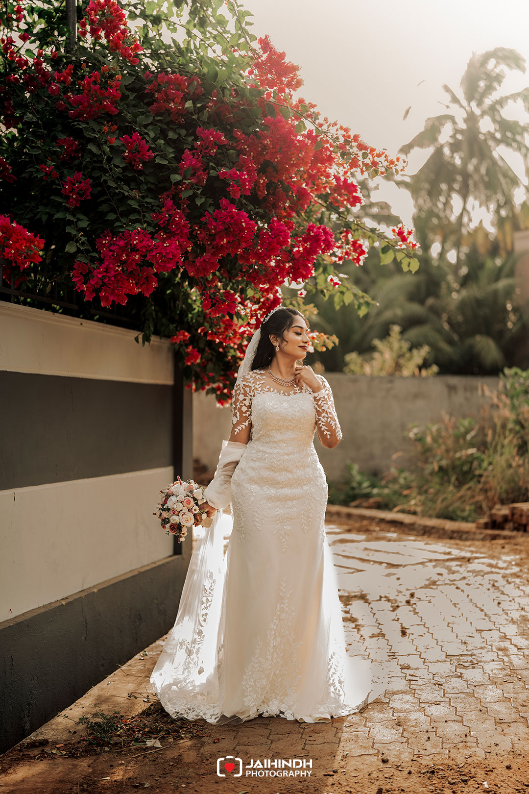 Bride in a white lace wedding gown holding a bouquet under a canopy of bright red bougainvillea flowers.