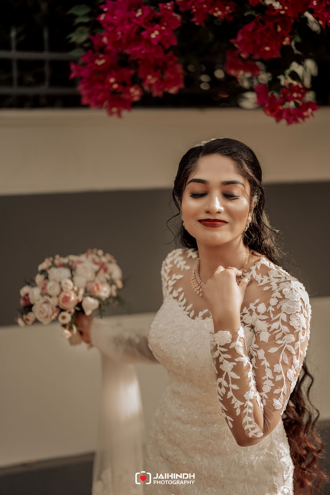 Bride in a lace wedding gown holding a bouquet, with eyes closed beneath hanging pink flowers.