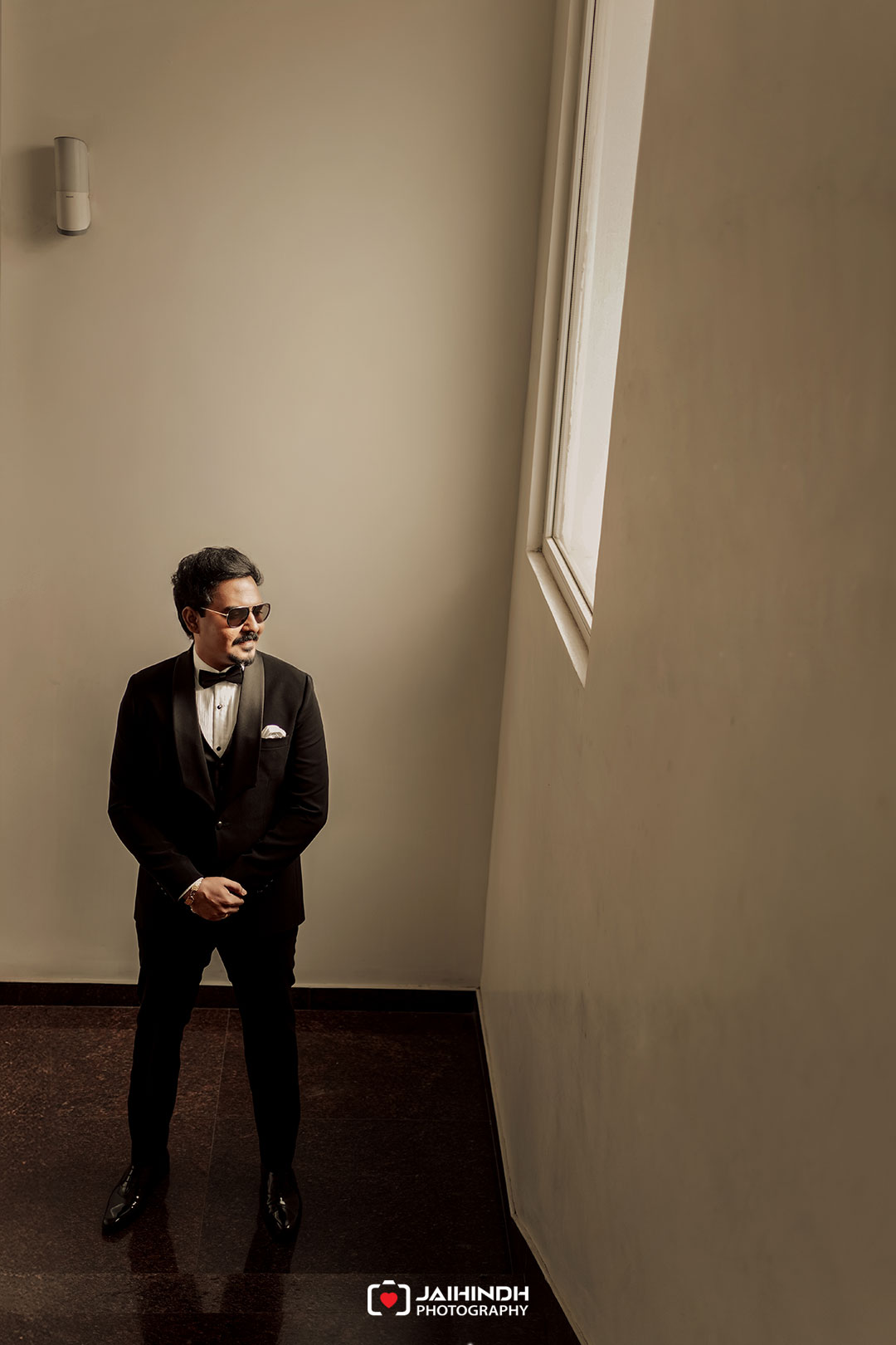 Man in a black tuxedo with bow tie and sunglasses stands with hands clasped in a sunlit, minimalist hallway by a large window.