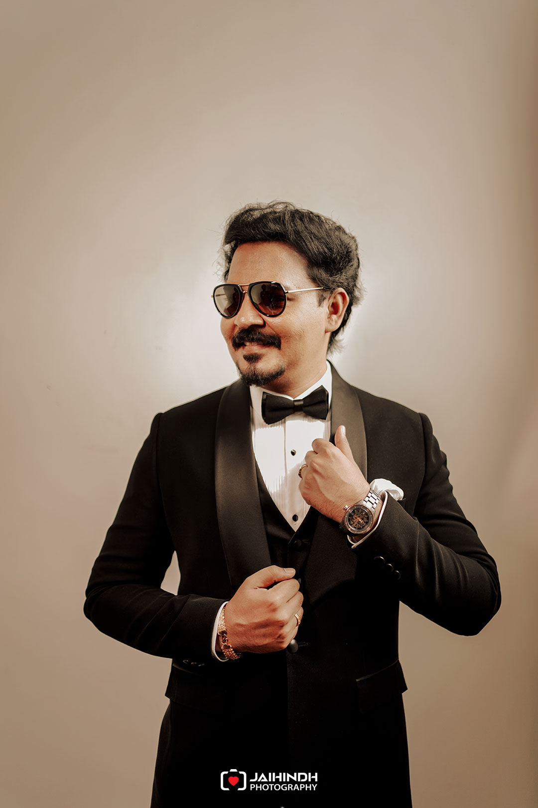 Man in a black tuxedo with bow tie, wearing sunglasses, posing confidently for a formal portrait.