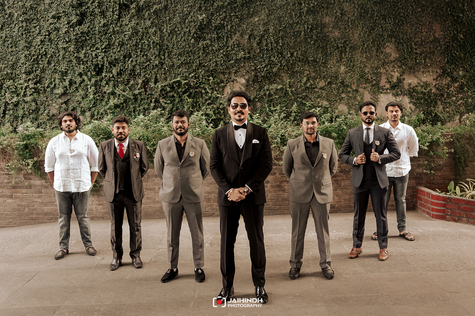 Groom in a black tuxedo with sunglasses stands center, flanked by six groomsmen in suits outdoors against an ivy-covered wall.