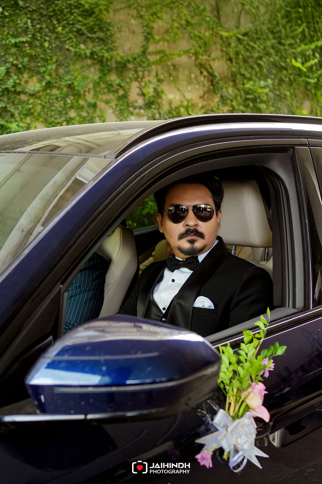 Man in a black tuxedo and sunglasses sits in a dark blue car, with a floral bouquet attached to the front, ready for a wedding scene.